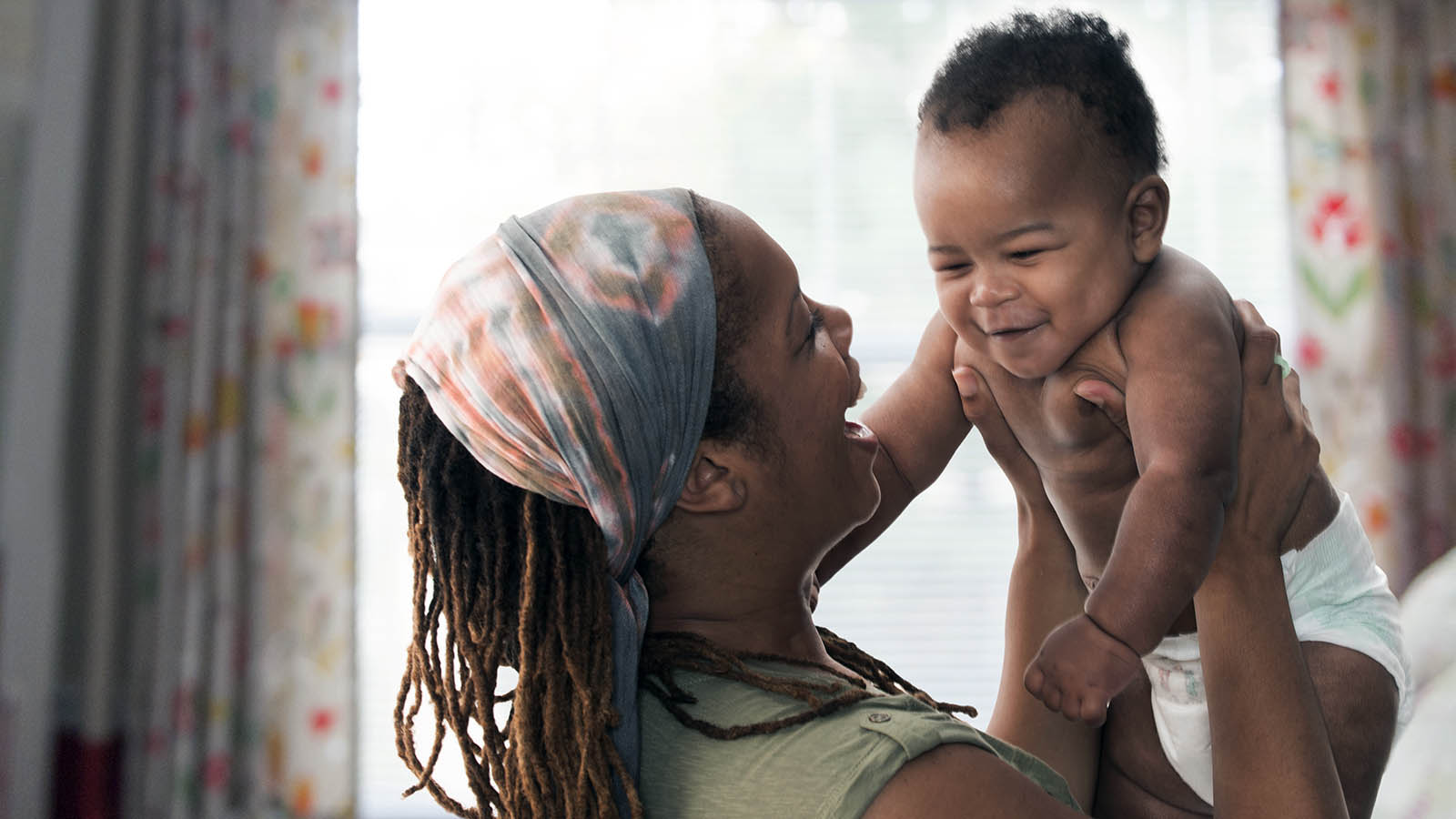 A mother holding her baby up, smiling — illustrative of parent–child interaction and the strain of lifting that can cause 'mommy thumb'.
